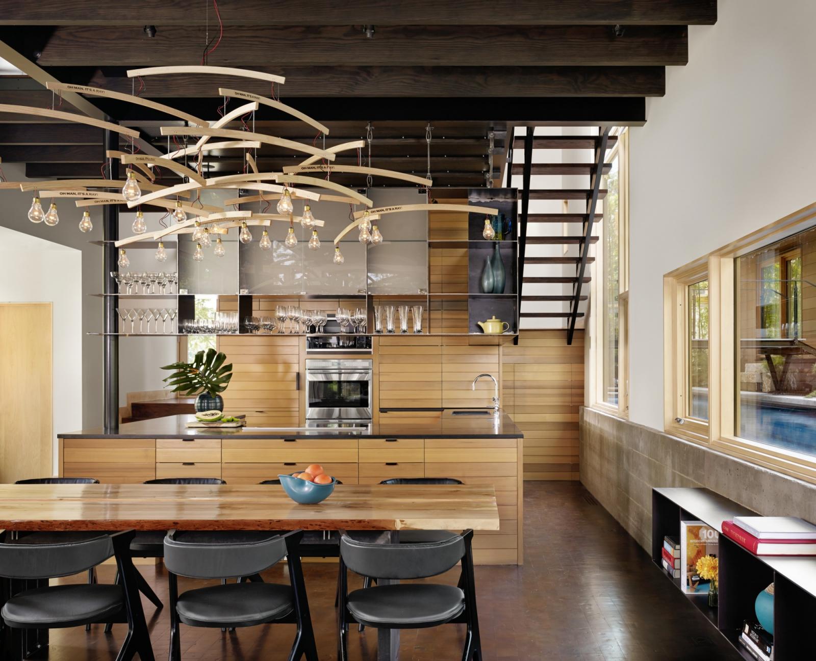 Looking across custom wood dining table and chandelier with view to open kitchen Situated at the confluence of Hog Pen Creek and Lake Austin, Hog Pen Creek Residence was envisioned by its owners as a place that evokes the playfulness of summer on the lake. Interior image looking across custom wood dining table and chandelier with view